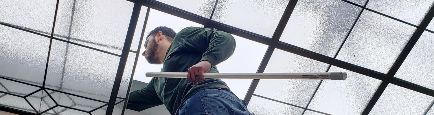 A photo showing a worker on a ladder changing out long light bulbs on the ceiling.