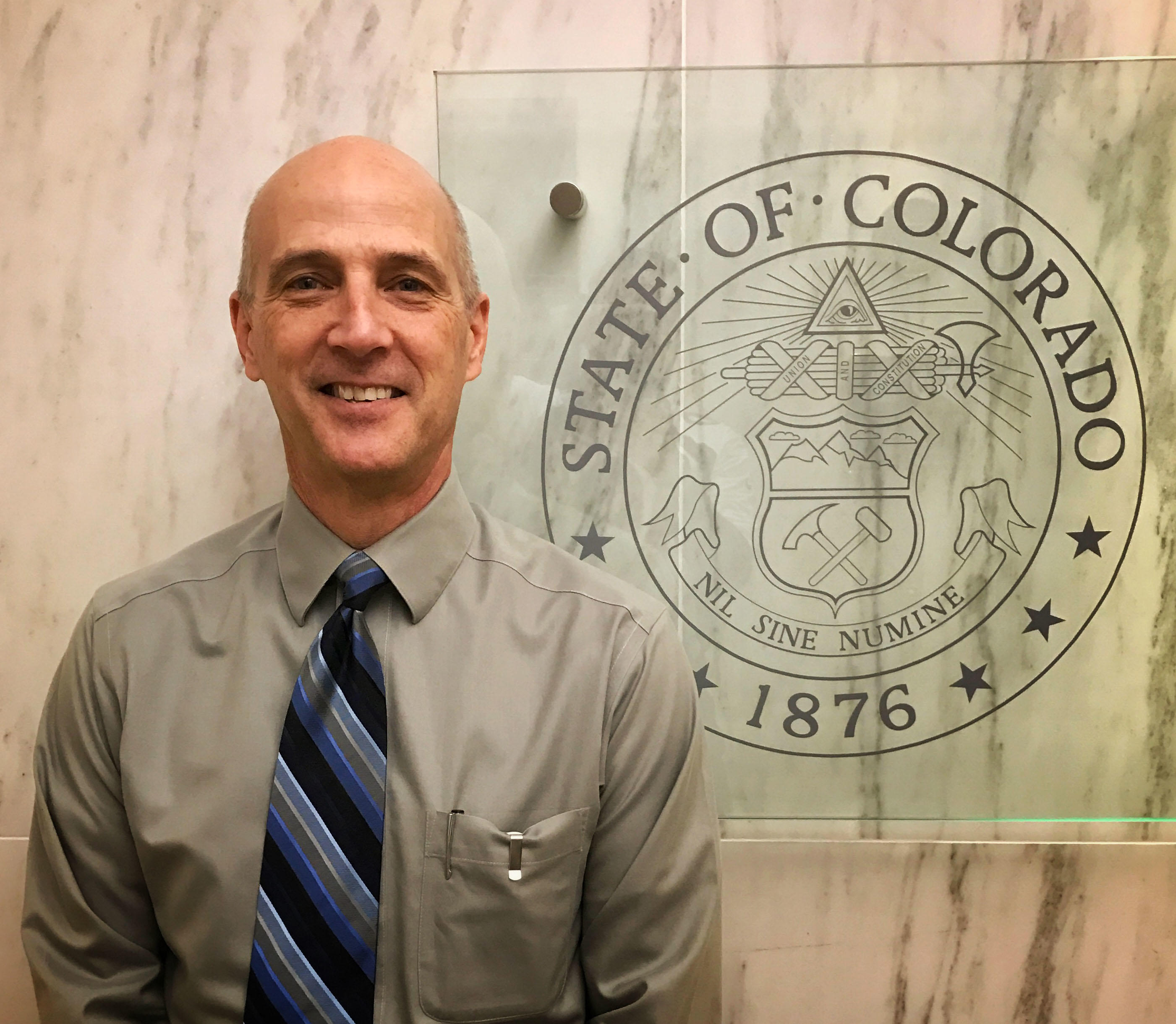 Photo of Rick Lee standing in front of the State of Colorado Seal