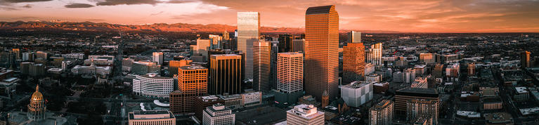 A panoramic photo of the city of Denver with an orange sunset vibe with the State Capitol Dome shining in the lower left corner and other buildings showing including the mailbox building, etc.